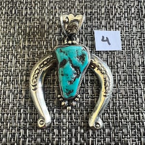 May include: A silver pendant featuring a central triangular turquoise stone with dark matrix veins. The silver frame is shaped like an inverted horseshoe with engraved floral patterns and a decorative bail. A small white tag with the number 4 is visible.