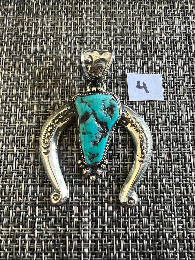 May include: A silver pendant featuring a central triangular turquoise stone with dark matrix veins. The silver frame is shaped like an inverted horseshoe with engraved floral patterns and a decorative bail. A small white tag with the number 4 is visible.