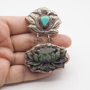 May include: A silver pendant featuring a carved green lotus flower with a turquoise stone in the center. The pendant is attached to a silver chain with a decorative lotus flower design.