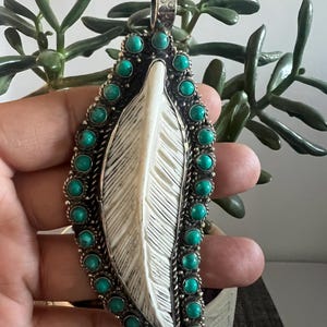 May include: A large pendant featuring a central carved white feather-shaped element surrounded by a border of small, round, bright turquoise stones set in a dark, oxidized silver frame with a textured metal bail at the top.