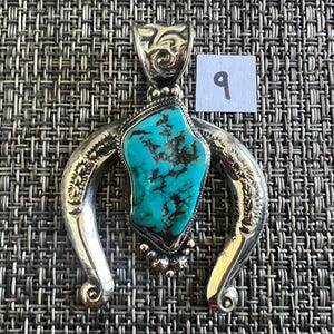 May include: A silver pendant featuring a central irregular turquoise stone with dark matrix veins, set within a horseshoe-shaped frame with engraved floral details and a decorative bail, accompanied by a small white square label with the number 9.