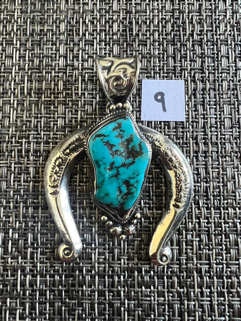 May include: A silver pendant featuring a central irregular turquoise stone with dark matrix veins, set within a horseshoe-shaped frame with engraved floral details and a decorative bail, accompanied by a small white square label with the number 9.