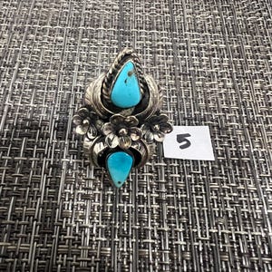 May include: A silver ring featuring two teardrop-shaped turquoise stones, one at the top and one at the bottom, surrounded by metal floral and leaf motifs. A small white square tag with the handwritten number 5 is placed next to the ring.