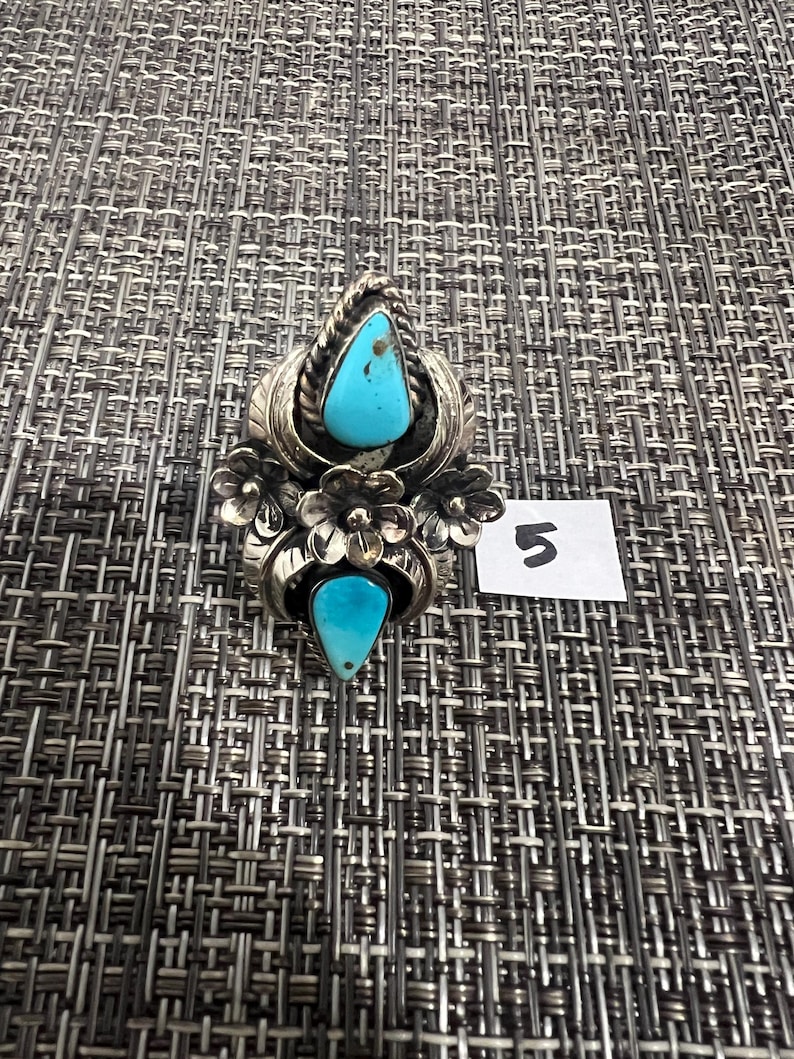 May include: A silver ring featuring two teardrop-shaped turquoise stones, one at the top and one at the bottom, surrounded by metal floral and leaf motifs. A small white square tag with the handwritten number 5 is placed next to the ring.