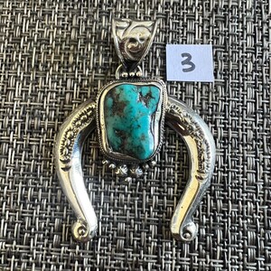 May include: A silver pendant featuring a central, irregular turquoise stone with dark matrix veins, set within a horseshoe-shaped frame with engraved leaf-like patterns and a decorative bail. A small white tag with the number 3 is attached.