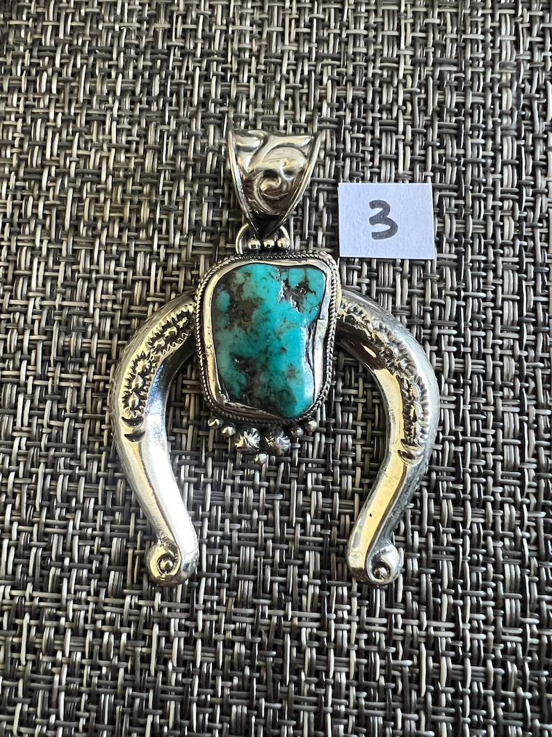 May include: A silver pendant featuring a central, irregular turquoise stone with dark matrix veins, set within a horseshoe-shaped frame with engraved leaf-like patterns and a decorative bail. A small white tag with the number 3 is attached.