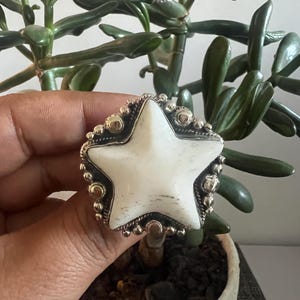 May include: A star-shaped ring featuring a smooth white stone centerpiece set in a dark metal frame. The setting is decorated with small, round metal beads along the edges, creating a textured border around the central white star shape.