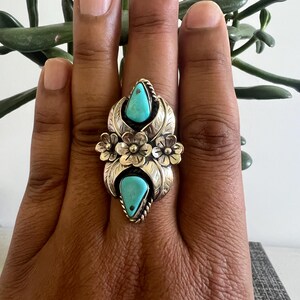 May include: A silver ring featuring two teardrop-shaped turquoise stones set vertically. The metalwork includes three dimensional silver flowers in the center and leaf-like engravings surrounding the stones, with twisted wire detailing around the bezels.