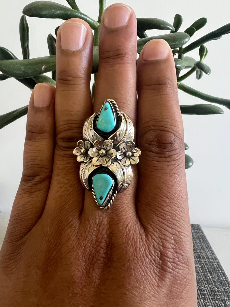 May include: A silver ring featuring two teardrop-shaped turquoise stones set vertically. The metalwork includes three dimensional silver flowers in the center and leaf-like engravings surrounding the stones, with twisted wire detailing around the bezels.