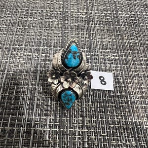 May include: A silver ring featuring two teardrop-shaped turquoise stones with dark matrix patterns, accented by three dimensional silver flowers and leaf motifs. A small white tag with the number 8 is positioned next to the ring.