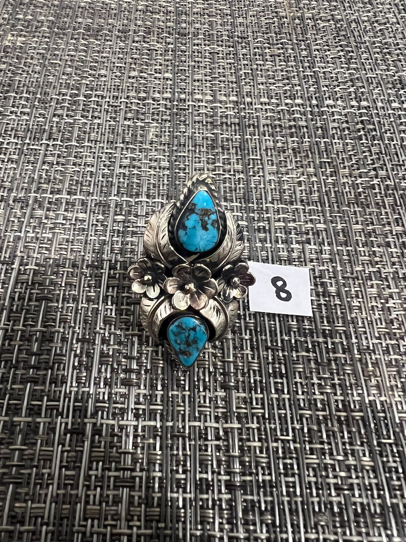 May include: A silver ring featuring two teardrop-shaped turquoise stones with dark matrix patterns, accented by three dimensional silver flowers and leaf motifs. A small white tag with the number 8 is positioned next to the ring.