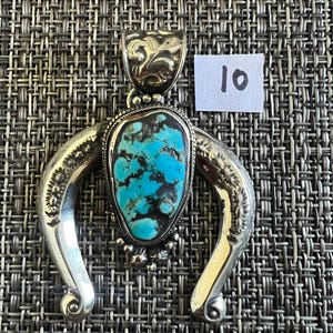 May include: A silver-toned pendant featuring a central teardrop-shaped turquoise stone with dark matrix veining. The metal frame has a curved, horseshoe-like shape with engraved floral details and a decorative bail. A small white tag displays the number 10.