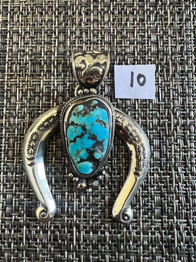 May include: A silver-toned pendant featuring a central teardrop-shaped turquoise stone with dark matrix veining. The metal frame has a curved, horseshoe-like shape with engraved floral details and a decorative bail. A small white tag displays the number 10.