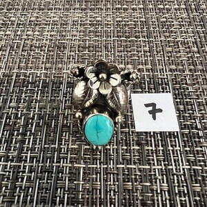 May include: A silver ring featuring a central oval turquoise stone with dark veining, topped with a detailed metal flower and leaf design. A small white square tag with the handwritten number 7 is positioned next to the ring.