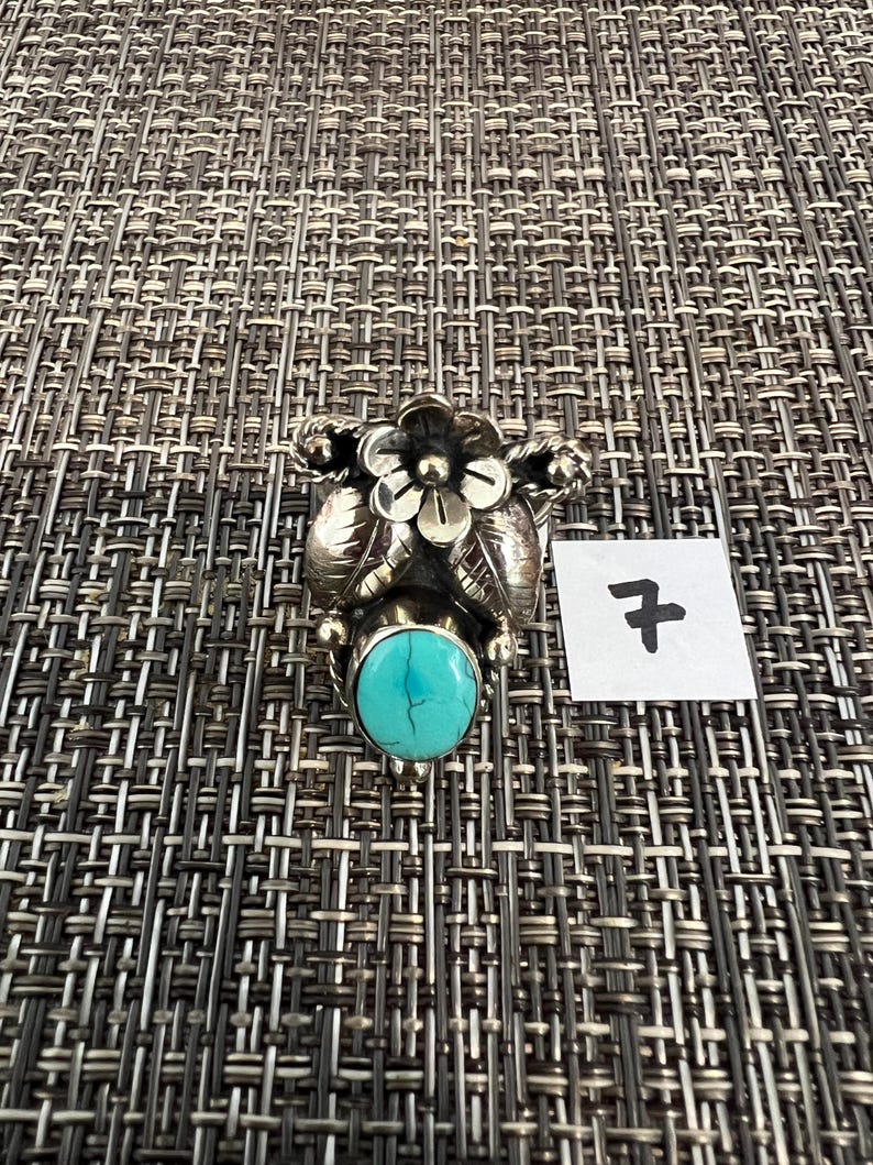 May include: A silver ring featuring a central oval turquoise stone with dark veining, topped with a detailed metal flower and leaf design. A small white square tag with the handwritten number 7 is positioned next to the ring.