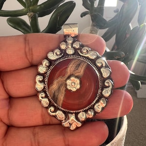 May include: A silver pendant with a round, red and cream-colored stone center. The stone has a floral design in the center. The pendant has an ornate, swirling silver border. The pendant is held in a hand.
