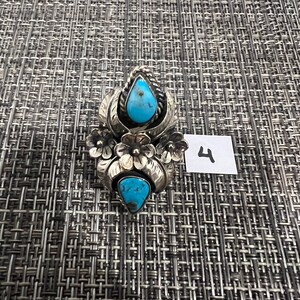 May include: A silver ring featuring two teardrop-shaped turquoise stones set vertically. The metalwork includes floral motifs with engraved leaves and petals surrounding the stones. A small white square tag with the handwritten number 4 is present.