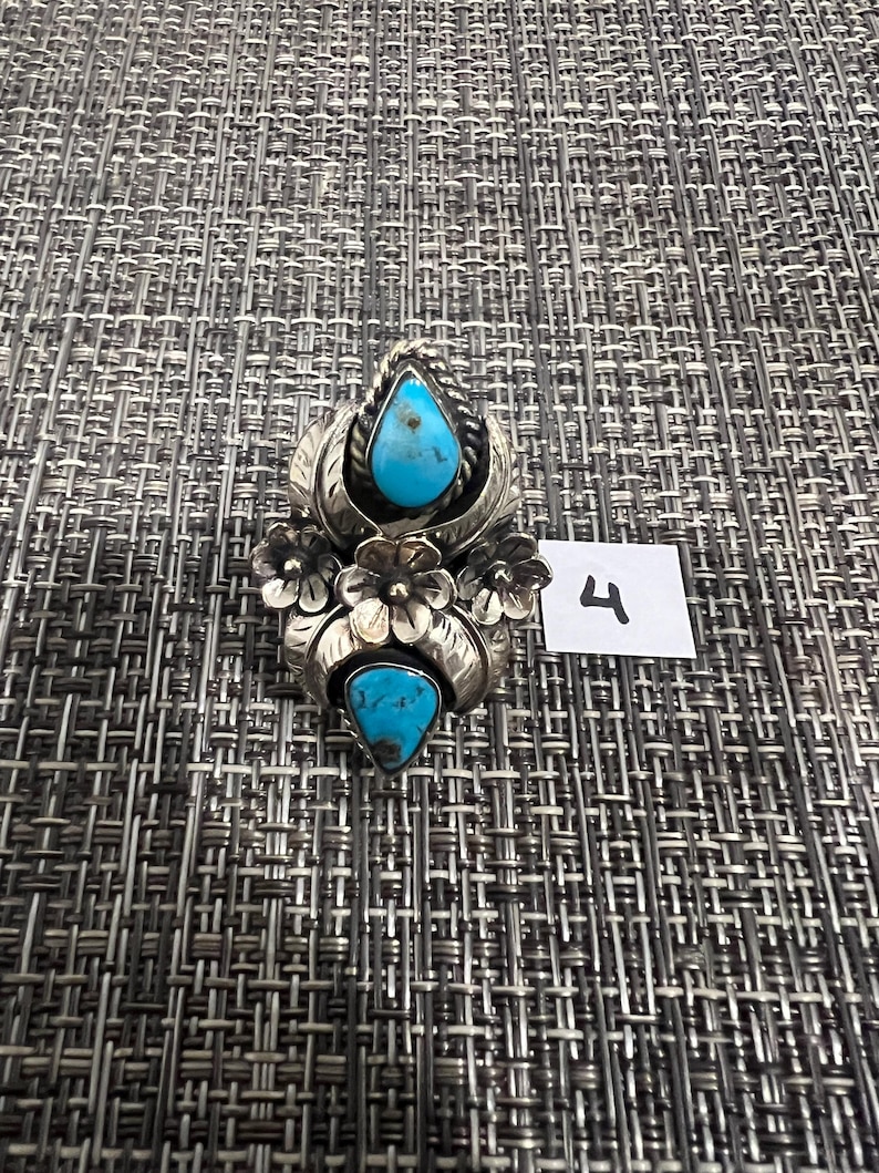 May include: A silver ring featuring two teardrop-shaped turquoise stones set vertically. The metalwork includes floral motifs with engraved leaves and petals surrounding the stones. A small white square tag with the handwritten number 4 is present.