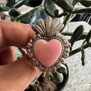 Op de afbeelding: Een zilverkleurige ring met een roze hartvormige steen. De ring heeft een sierlijk, gedetailleerd ontwerp rond het hart en een decoratieve bovenkant. De ring wordt vastgehouden voor een achtergrond van groene planten.