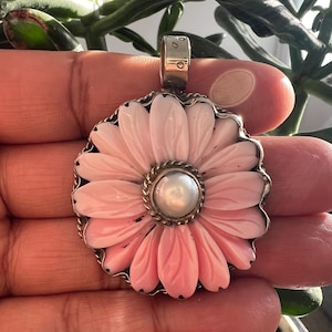 May include: A circular pendant featuring a carved light pink flower with elongated petals and a central white pearl. The floral design is set within a silver frame with a twisted rope border and a silver bail at the top for attachment to a chain.