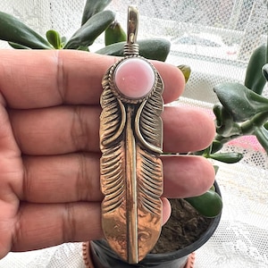 May include: A bronze-colored feather pendant with a pink gemstone at the top. The pendant has detailed feather etching and a silver-colored bail. The gemstone is a light pink color and is set in a silver-colored bezel.