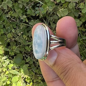 Op de afbeelding: Een zilveren ring met een grote ovale blauwe en witte edelsteen. De ring heeft een eenvoudige band met drie zilveren banden rond de edelsteen.