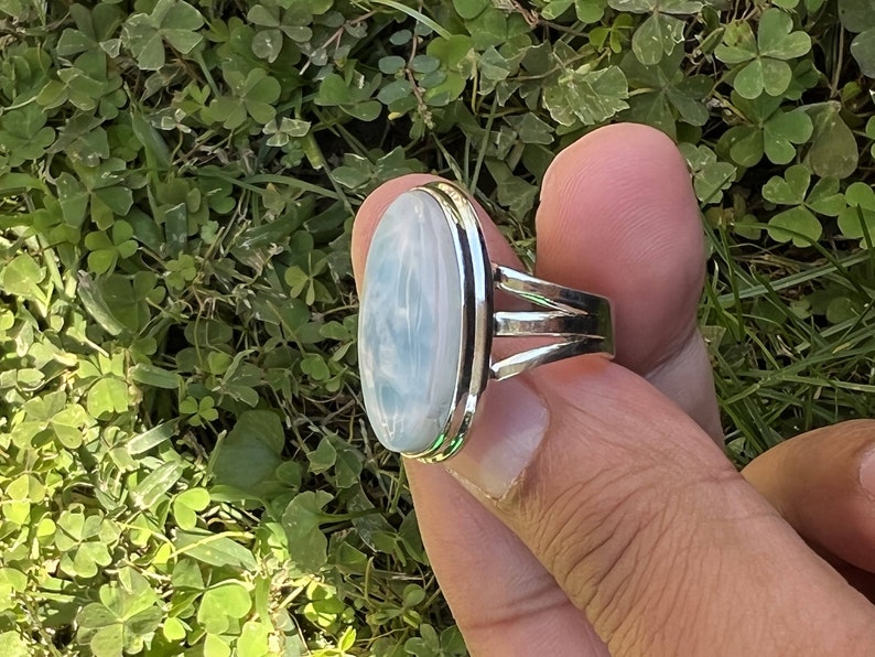 Op de afbeelding: Een zilveren ring met een grote ovale blauwe en witte edelsteen. De ring heeft een eenvoudige band met drie zilveren banden rond de edelsteen.