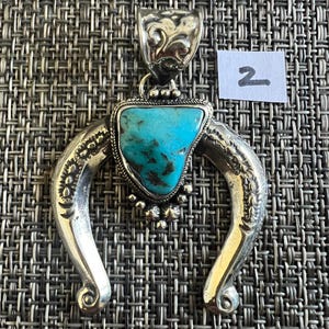 May include: A silver pendant featuring a triangular turquoise stone with dark matrix, set within a horseshoe-shaped frame with engraved patterns and a decorative bail. A small white paper tag with the handwritten number 2 is visible nearby.