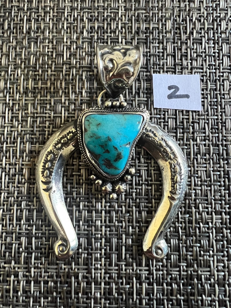May include: A silver pendant featuring a triangular turquoise stone with dark matrix, set within a horseshoe-shaped frame with engraved patterns and a decorative bail. A small white paper tag with the handwritten number 2 is visible nearby.