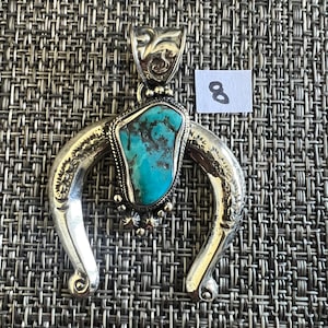 May include: A silver-toned horseshoe-shaped pendant featuring a central irregular turquoise stone with dark matrix veins. The metal frame has engraved floral details and a decorative bail. A small white paper tag with the number 8 is visible.