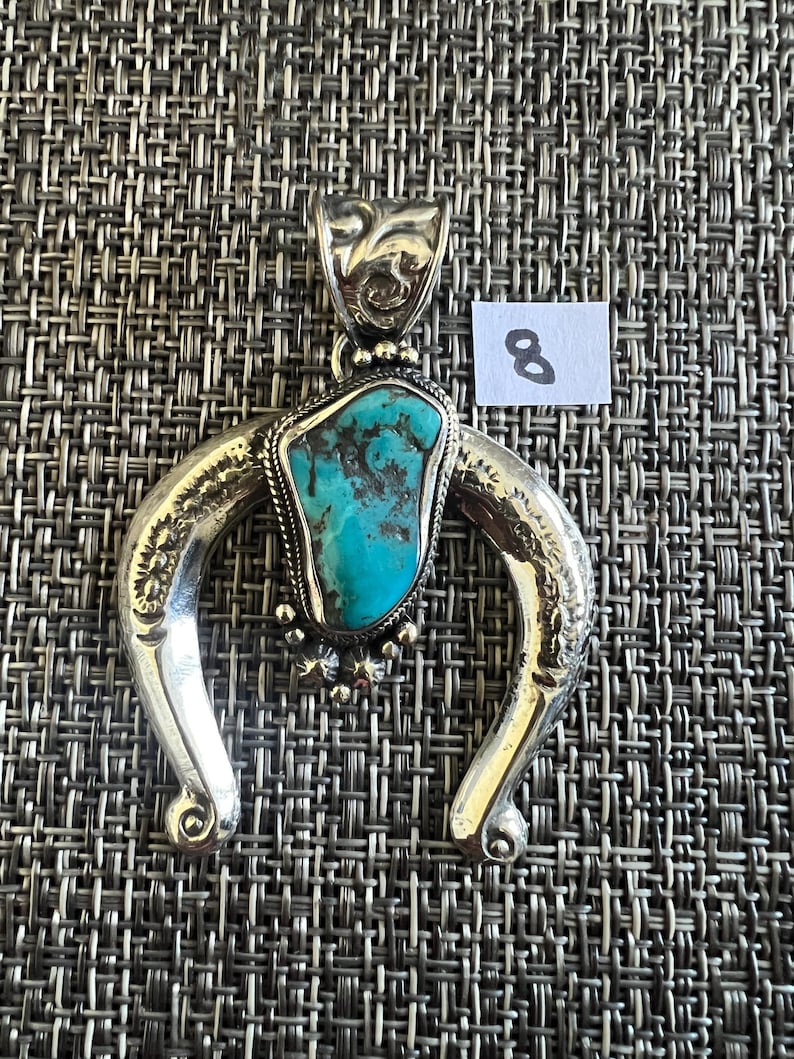 May include: A silver-toned horseshoe-shaped pendant featuring a central irregular turquoise stone with dark matrix veins. The metal frame has engraved floral details and a decorative bail. A small white paper tag with the number 8 is visible.