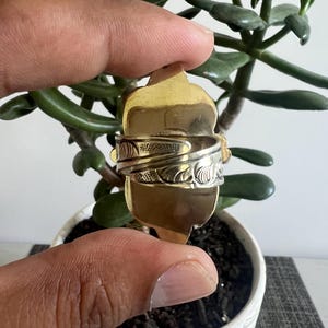 May include: A gold-toned ring featuring an elongated, shield-shaped face and a wide, adjustable band. The band displays a detailed, embossed floral or leaf-like pattern across its surface, while the central face remains smooth and polished.