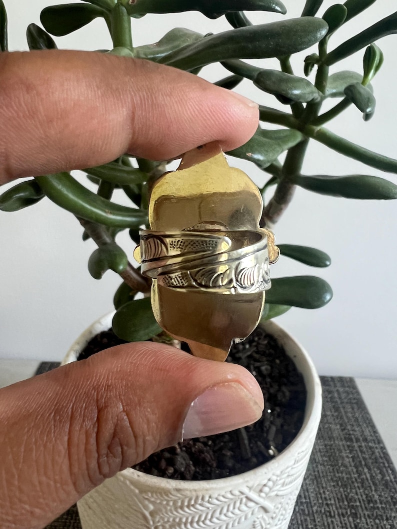 May include: A gold-toned ring featuring an elongated, shield-shaped face and a wide, adjustable band. The band displays a detailed, embossed floral or leaf-like pattern across its surface, while the central face remains smooth and polished.