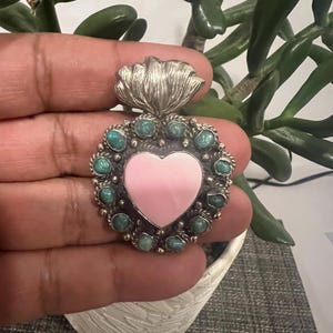 May include: A heart-shaped pendant crafted from silver, featuring a smooth, light pink center stone surrounded by a border of small, round, green turquoise cabochons. The top of the pendant is adorned with a stylized, flame-like silver element.