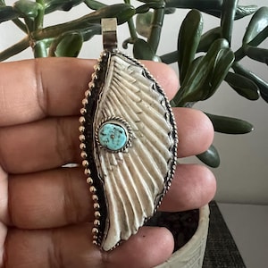 May include: A wing-shaped pendant featuring a carved off-white material with a textured surface, accented by a small, round, bright blue turquoise stone in the centre. The piece is framed in silver metal with a beaded edge and a top bail.