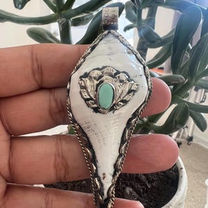 May include: A handcrafted pendant featuring a white shell design with a turquoise stone center. The pendant is framed with a silver-toned metal and has a decorative bail. The pendant is held in a hand, with a plant in the background.