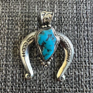 May include: A silver pendant featuring a crescent-shaped frame with engraved floral details and a central, irregular, bright blue turquoise stone with dark brown matrix veins, accented by a twisted wire border and small silver beads at the base.