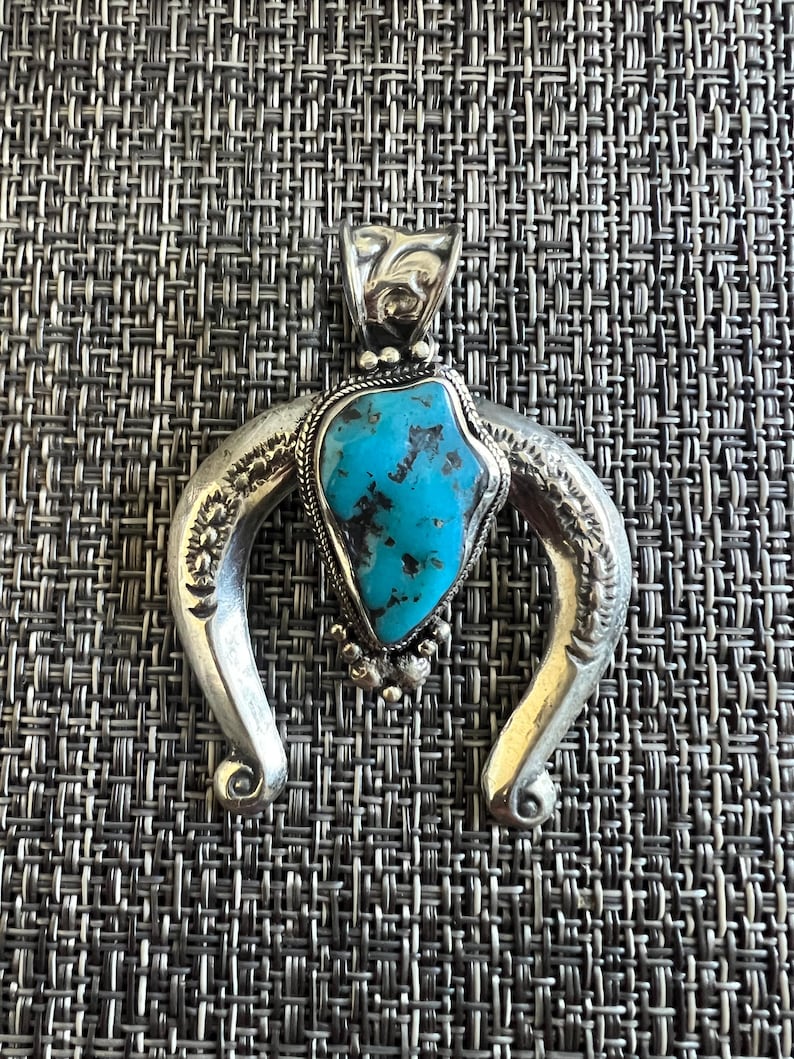 May include: A silver pendant featuring a crescent-shaped frame with engraved floral details and a central, irregular, bright blue turquoise stone with dark brown matrix veins, accented by a twisted wire border and small silver beads at the base.
