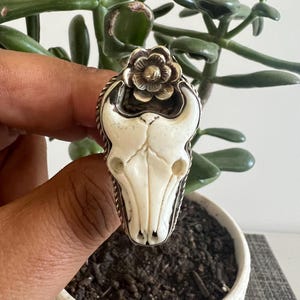 May include: A ring featuring a cream-colored carved bull skull centerpiece with a small silver flower accent at the top. The skull is set within a twisted silver metal border, creating a detailed focal point on the band of the ring.