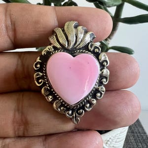May include: A heart-shaped brooch featuring a smooth, light pink center stone set within an ornate, dark silver-toned metal frame. The metalwork displays intricate scroll patterns and a flame-like design at the top, creating a detailed, layered appearance.