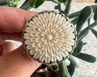 Op de afbeelding: Een crèmekleurige ring met een bloemmotief. De voorkant van de ring heeft een gedetailleerd, gesneden patroon dat op een bloem lijkt, gezet in een zilverkleurige metalen band. De ring wordt voor een groene plant gehouden.