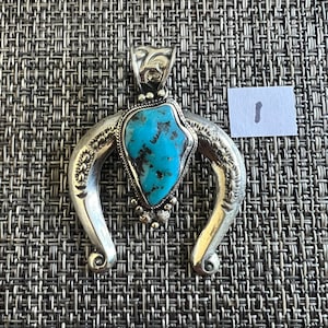 May include: A silver pendant featuring a central irregular turquoise stone with dark matrix, surrounded by a twisted wire border and small silver beads. The pendant has a curved, horseshoe-shaped frame with engraved details and a bail at the top.