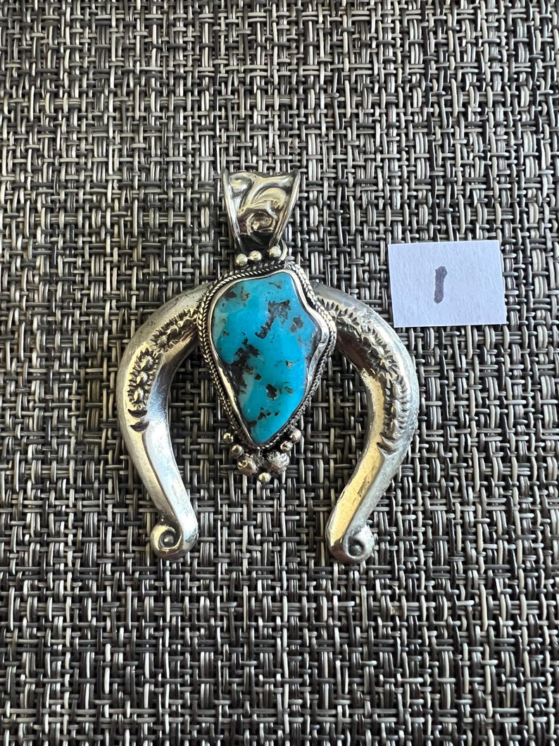 May include: A silver pendant featuring a central irregular turquoise stone with dark matrix, surrounded by a twisted wire border and small silver beads. The pendant has a curved, horseshoe-shaped frame with engraved details and a bail at the top.