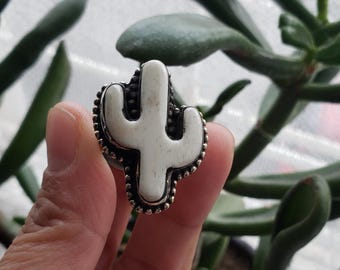 Op de afbeelding: Een zilverkleurige ring met een wit cactusontwerp. De cactus heeft een gedetailleerd, getextureerd oppervlak. De rand van de ring is versierd met kleine, parelachtige accenten. Groene bladeren op de achtergrond.