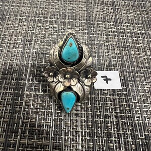 May include: A silver ring featuring two teardrop-shaped turquoise stones set vertically. The metalwork includes engraved leaf patterns and three small silver flowers between the stones. A small white tag with the handwritten number 7 is attached.