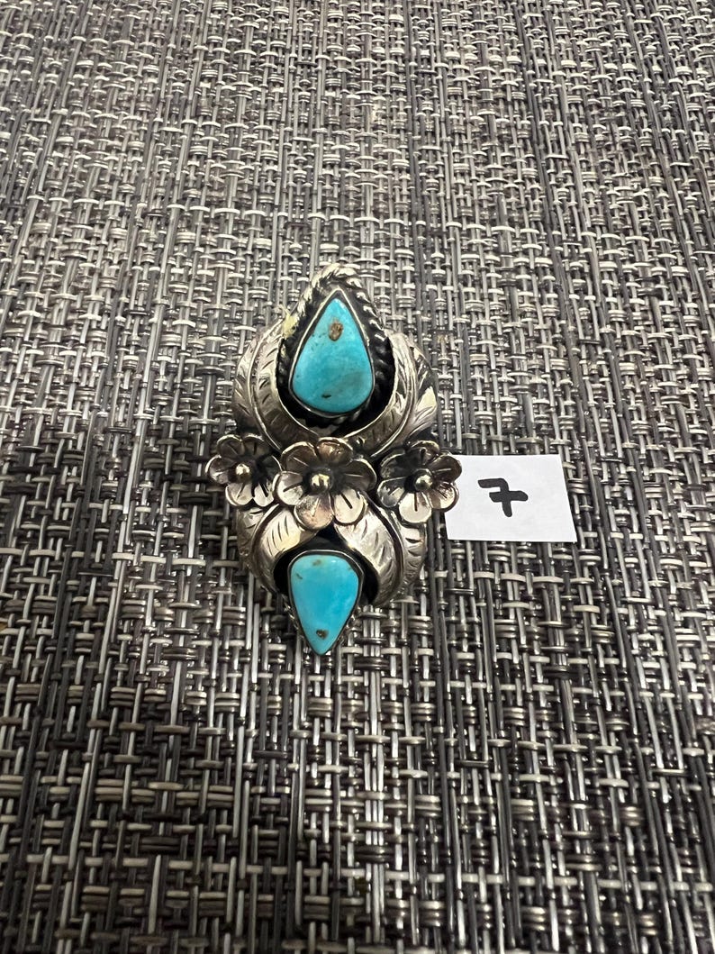 May include: A silver ring featuring two teardrop-shaped turquoise stones set vertically. The metalwork includes engraved leaf patterns and three small silver flowers between the stones. A small white tag with the handwritten number 7 is attached.