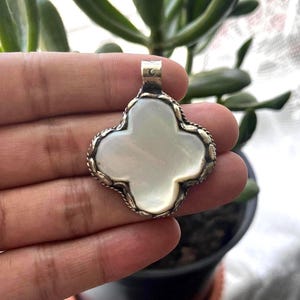 Puede incluir: Un colgante plateado con un diseño de trébol de cuatro hojas. El colgante presenta un centro blanco nacarado y un borde detallado en forma de cuerda retorcida. La fianza tiene una forma rectangular simple. El colgante es probablemente una joya.