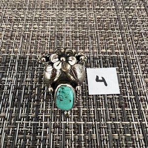 May include: A silver ring featuring a central oval turquoise stone with dark matrix inclusions, topped with a sculpted metal flower and leaf design. A small white square paper tag with the handwritten number 4 is placed next to the ring.