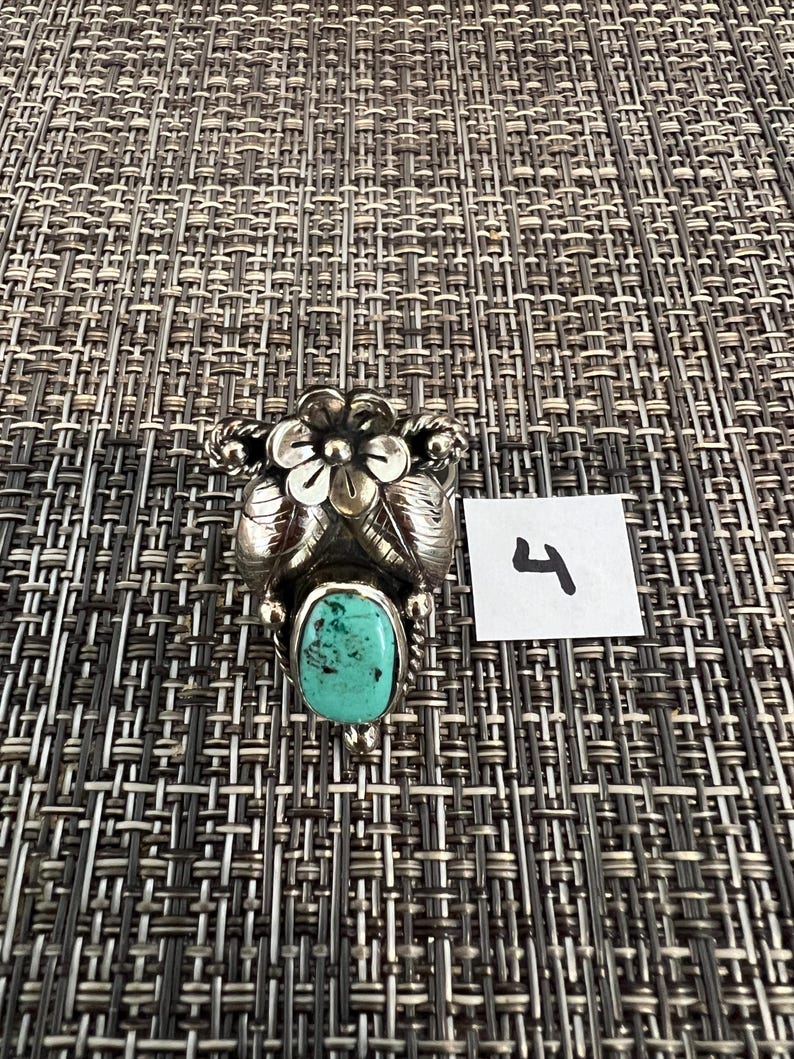 May include: A silver ring featuring a central oval turquoise stone with dark matrix inclusions, topped with a sculpted metal flower and leaf design. A small white square paper tag with the handwritten number 4 is placed next to the ring.