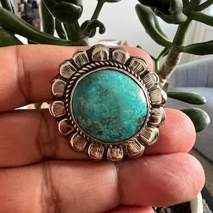 May include: A silver-toned ring with a large, round turquoise stone. The ring has a detailed, floral-like border. The turquoise stone is a vibrant teal color. The ring is held in a hand, with a green plant in the background.